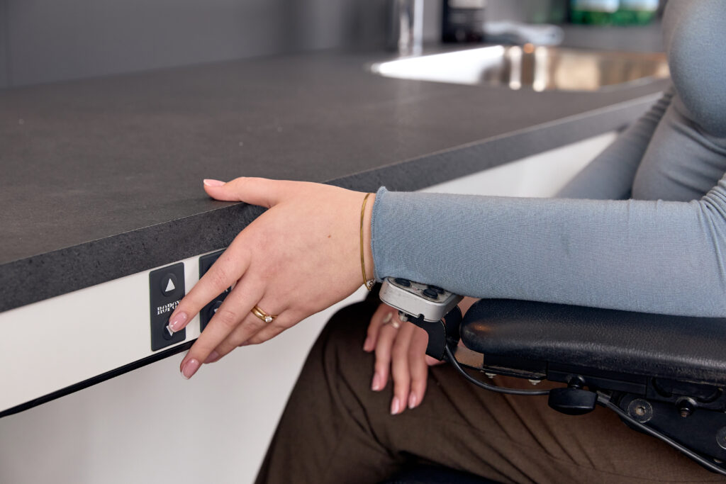 Wheelchair user pressing the Ropox control switch on the edge of a height-adjustable kitchen countertop to electrically adjust its height